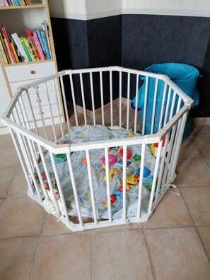 A white baby playpen with toys inside, a bookshelf with books, and a blue chair in the background.