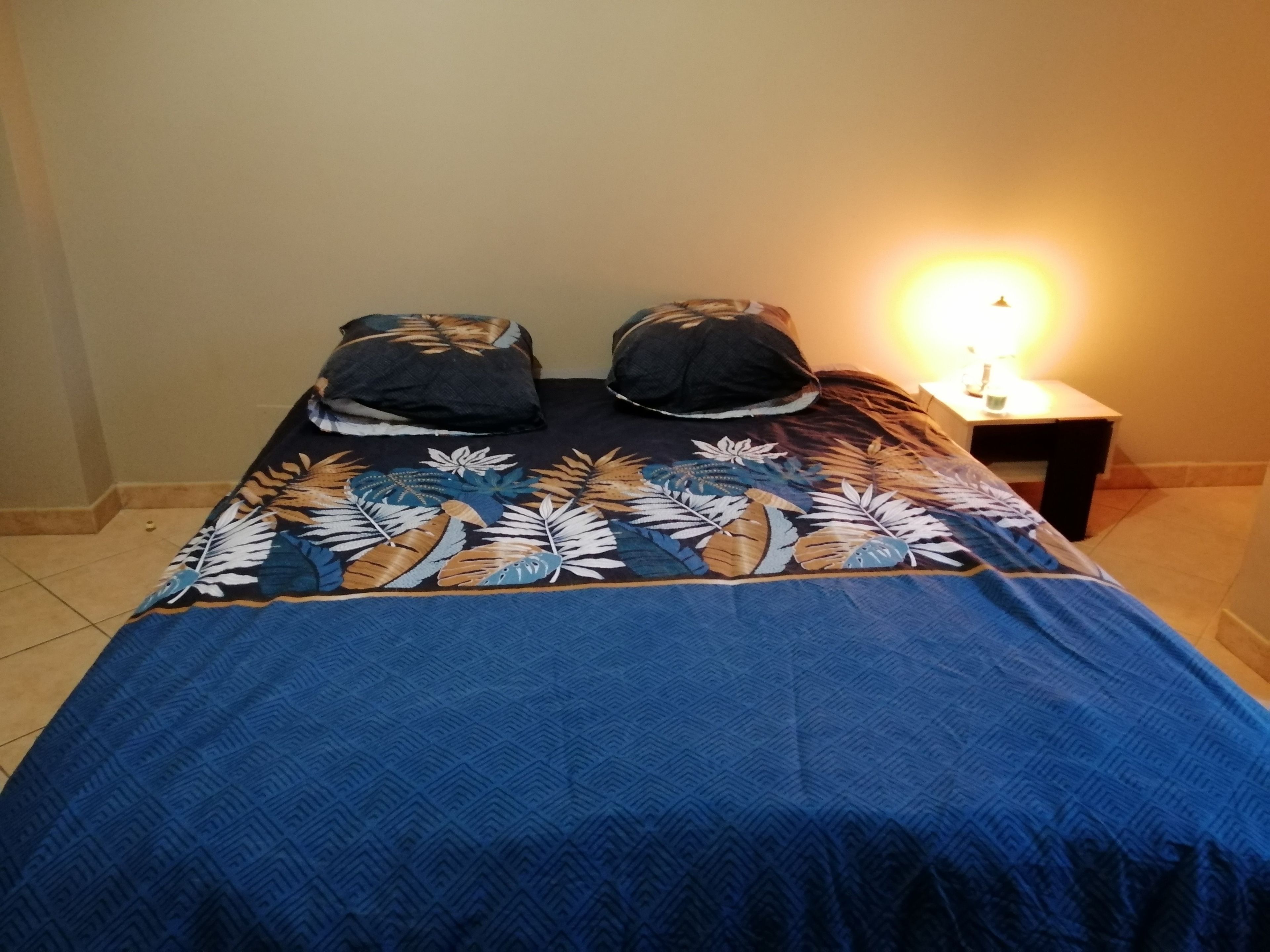 Een bed met een blauw bedovertrek met patroon en twee kussens, een nachtkastje met een lamp, en een betegelde vloer.