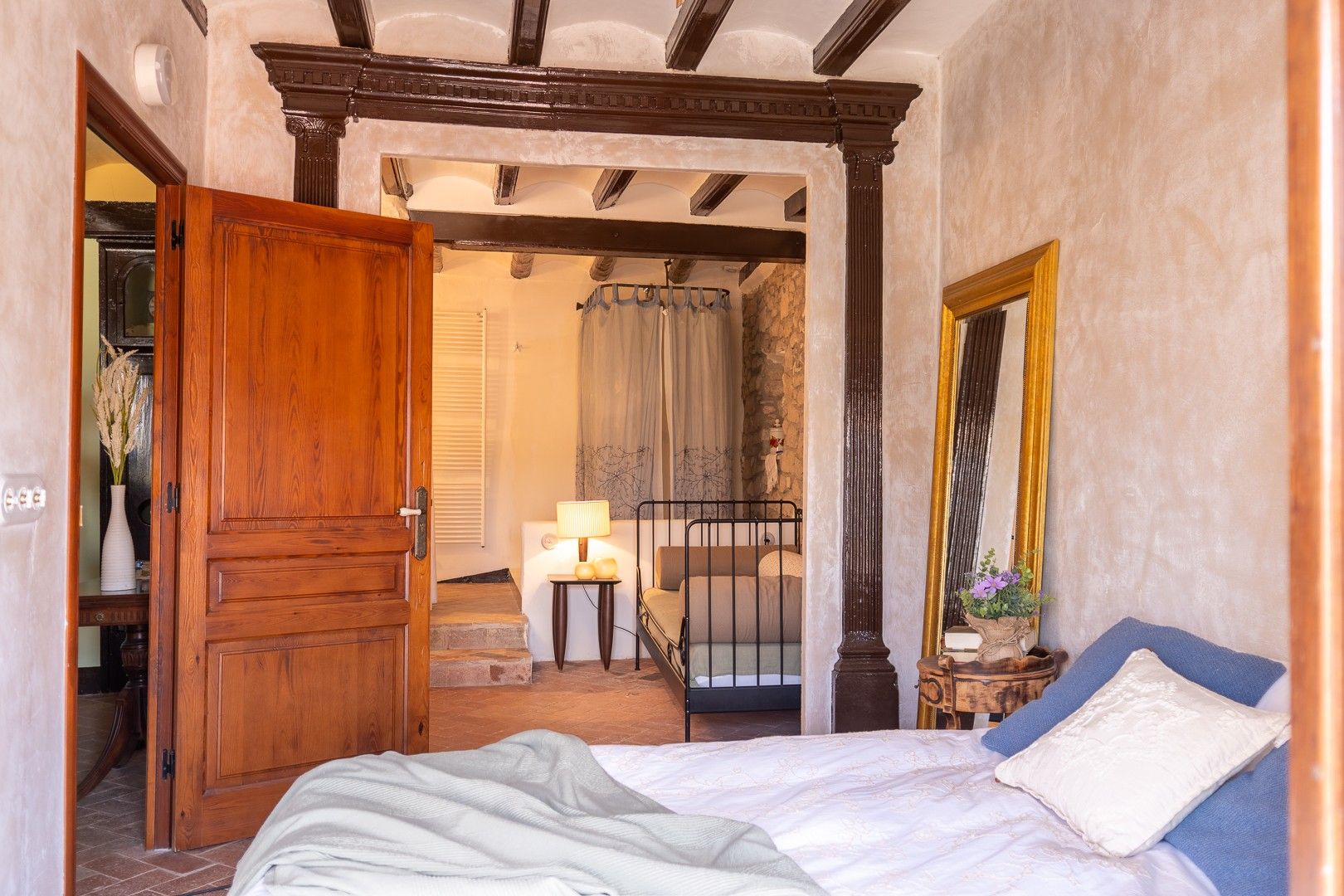 Een slaapkamer met een houten deur, een bed met kussens, een nachtkastje, een spiegel en een trap naar een badkamer.