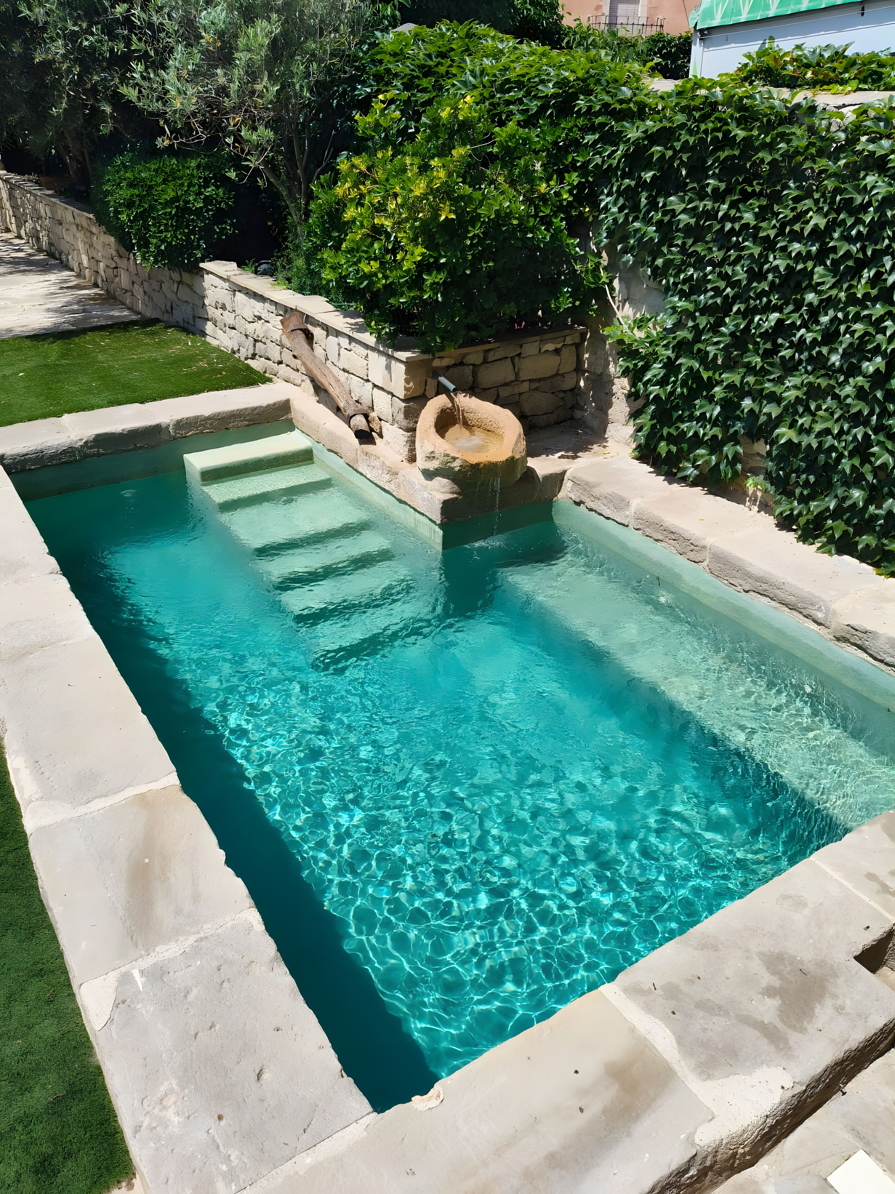 A rectangular swimming pool with clear blue water, surrounded by stone walls and lush greenery.