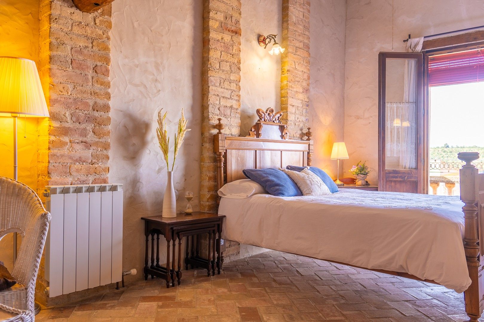 Un dormitorio con pared de ladrillo, cama con almohadas azules, mesita de noche con lámpara y ventana con vista.