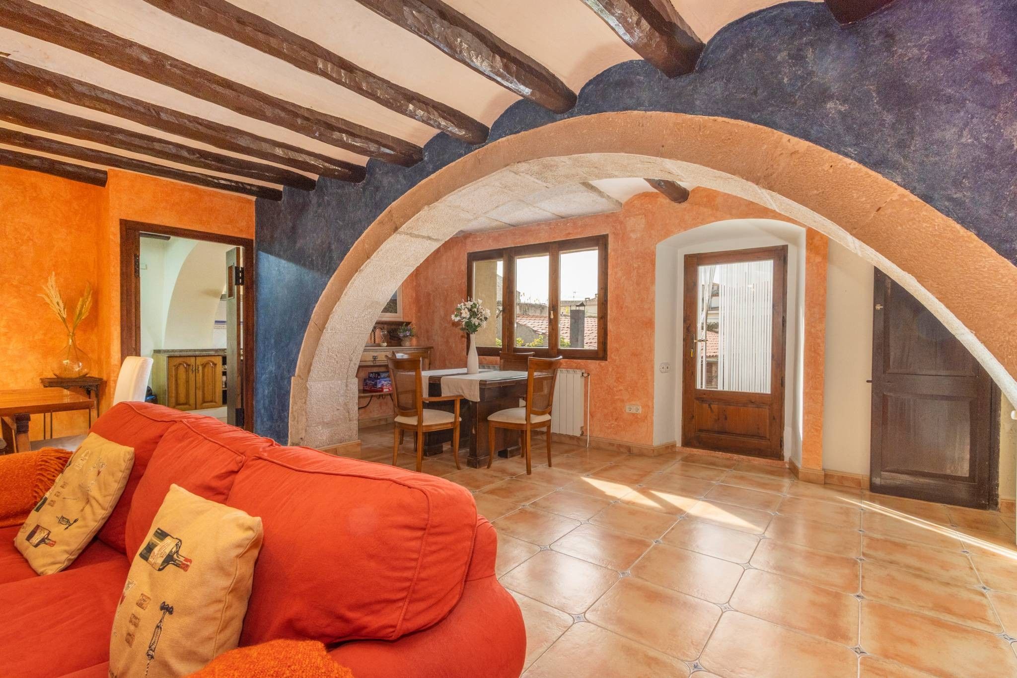 A cozy living room with a red sofa, an arched doorway leading to a dining area, and a tiled floor.