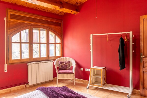A room with a red wall, a wooden-framed window, a wicker chair, a radiator, a small table, and a clothing rack. - Room 'La Vinya' with Kitchen Access, Garden, Pool and Wi-Fi (Sant Martí de Maldà)