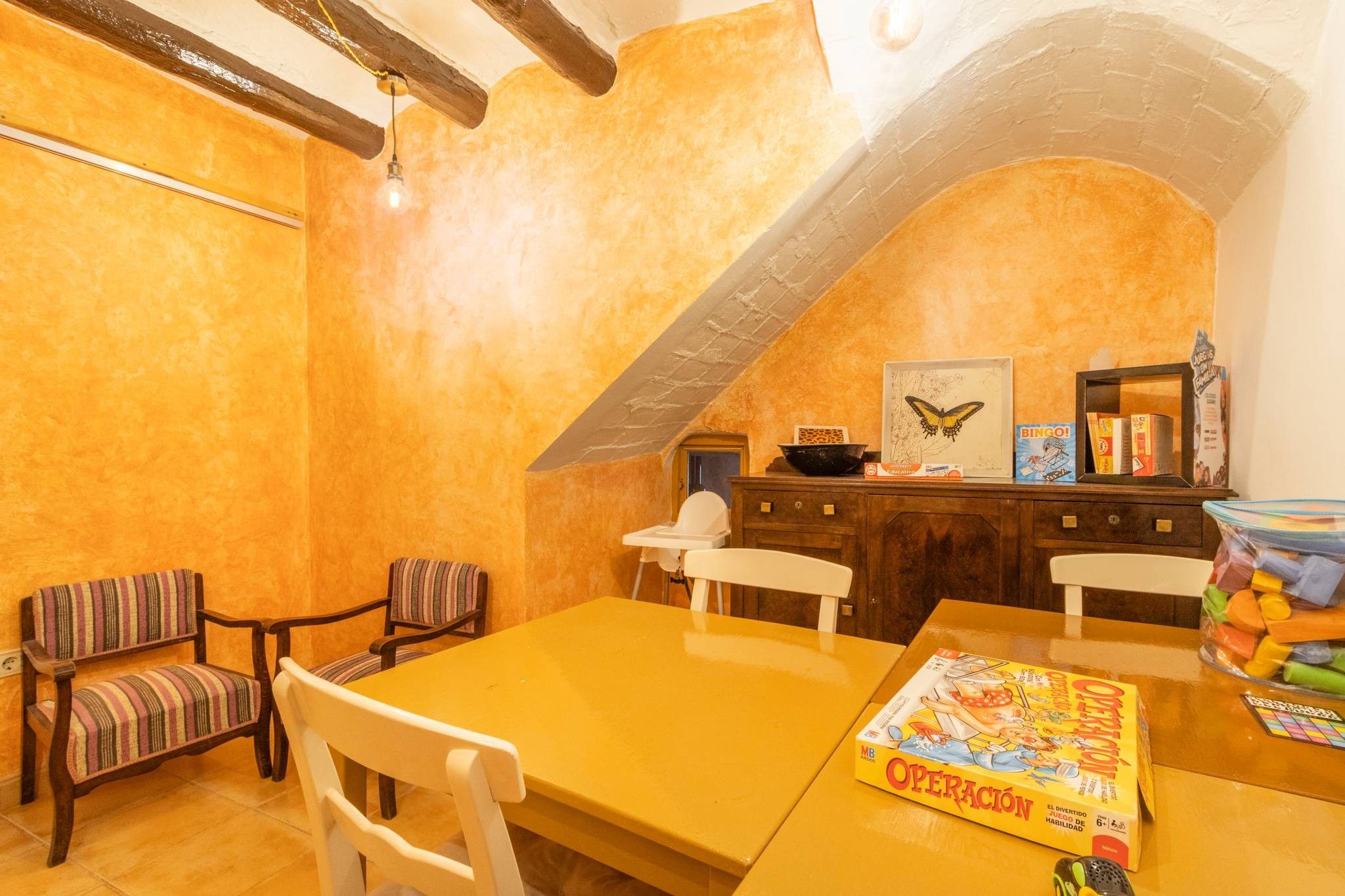 A dining area with a yellow table, two striped chairs, a white chair, and a bookshelf with various items including a BINGO game and a butterfly painting.