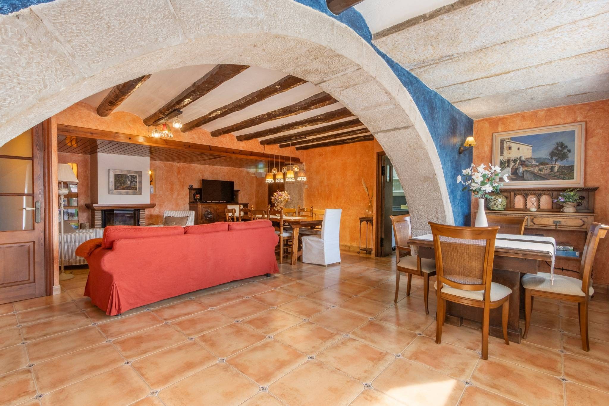 A spacious living area with a red sofa, wooden dining table, and chairs, under a vaulted ceiling with exposed beams.