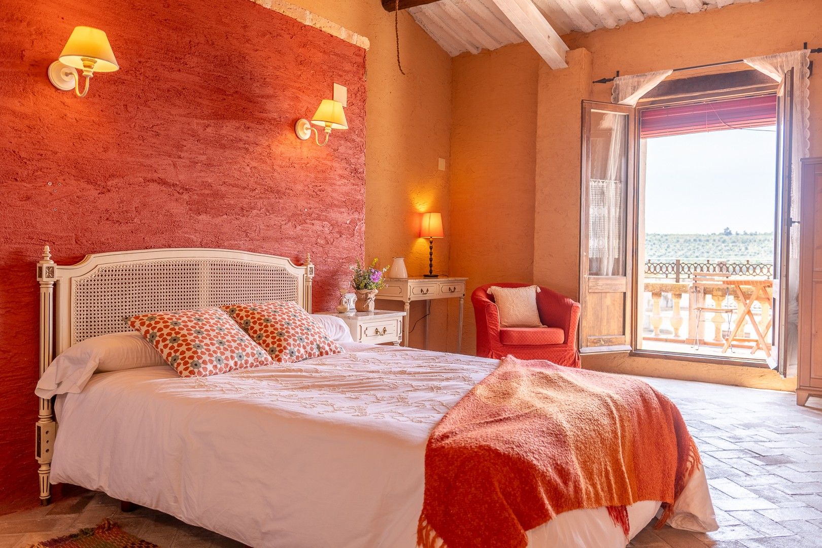 A bedroom with a bed, two wall-mounted lamps, a red chair, a small table with a lamp, and a view of the sea through an open door.
