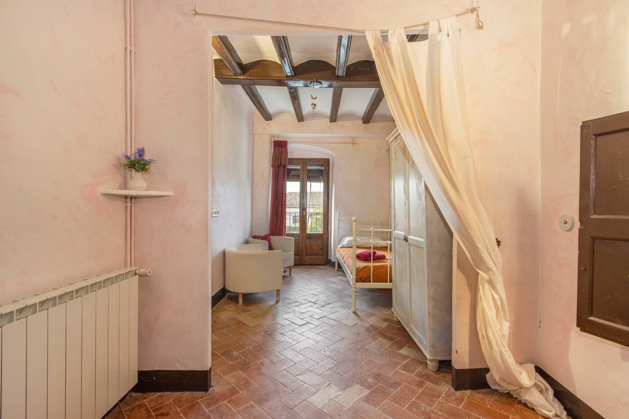 Un couloir étroit avec un radiateur, une étagère blanche sur laquelle est posé un vase, un lit avec une tête de lit et une fenêtre avec des rideaux.