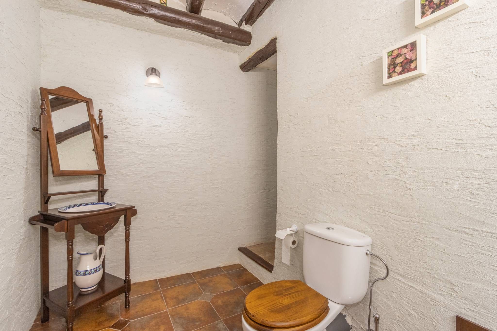 Un baño pequeño con inodoro de madera, un espejo, una repisa con jarrón y un dispensador de papel higiénico.
