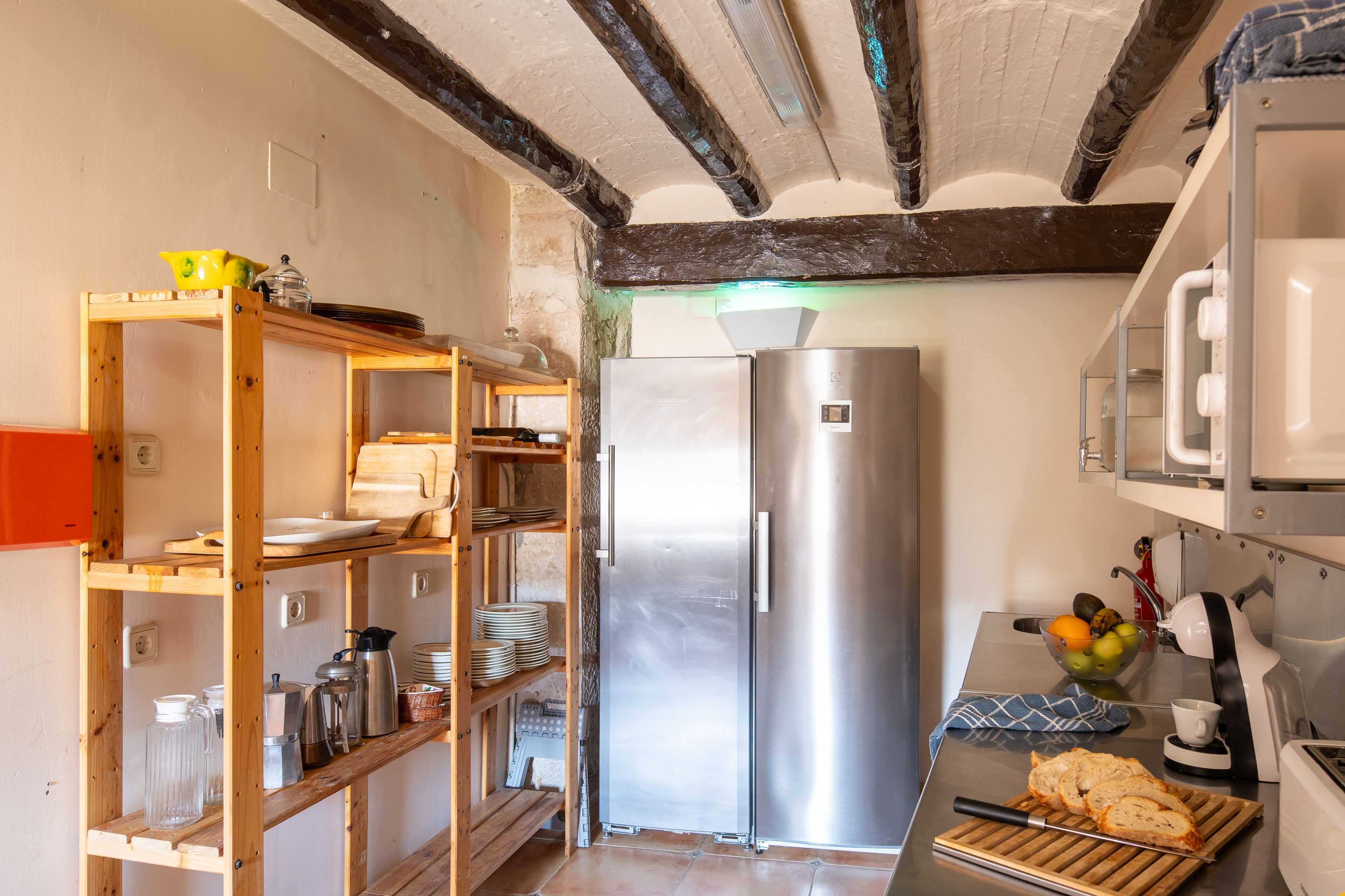 A compact kitchen with a built-in stainless steel refrigerator, wooden shelving, and a small countertop with a toaster and a fruit bowl.