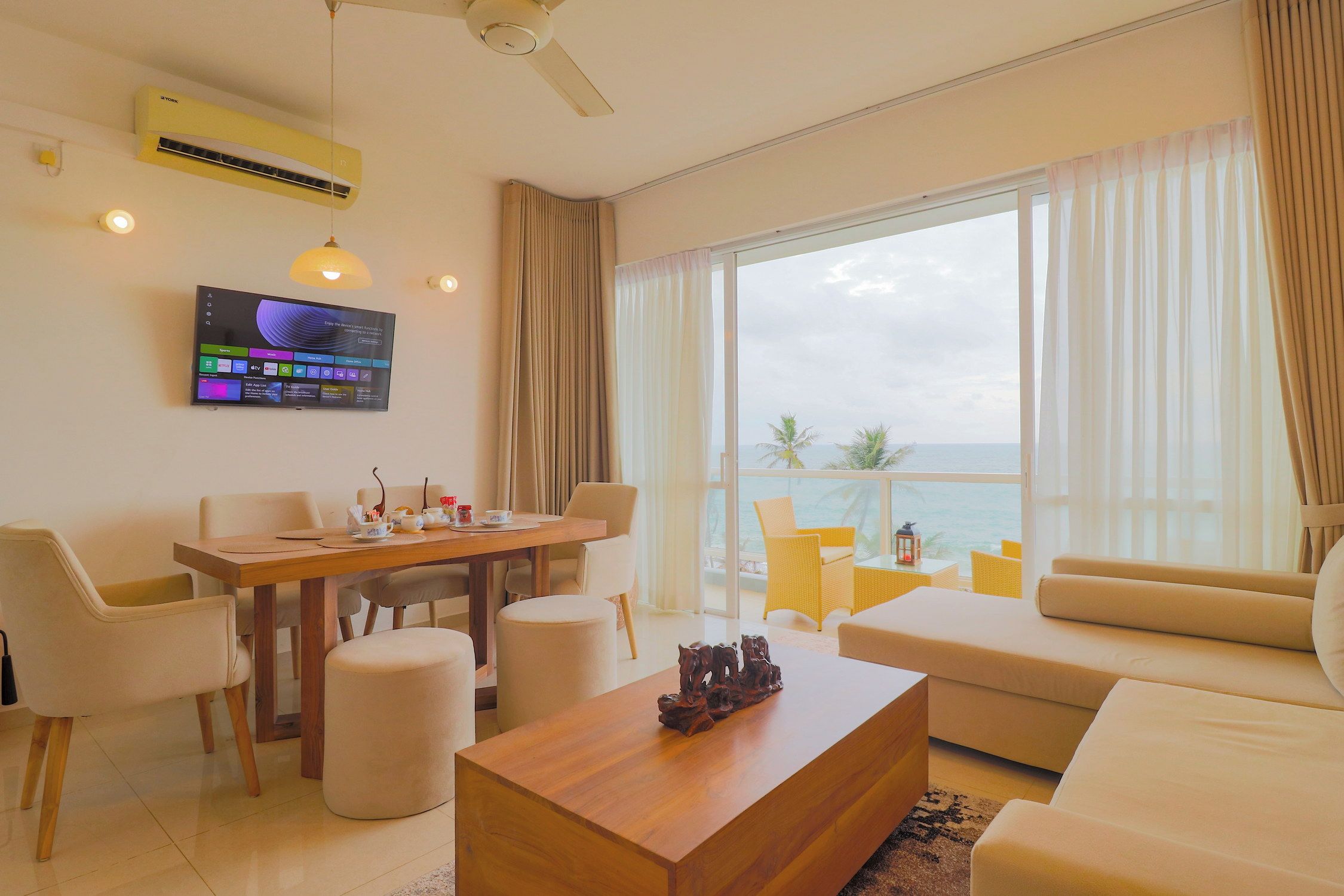 A modern living room with a flat-screen TV, a dining area, and a large window with a view of the ocean.