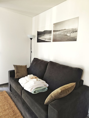 A living room with a dark grey sofa, a lamp, and two black and white beach photographs on the wall. - Tiny Home 'Casa Azul' with Sea View, Shared Pool and Wi-Fi (Cristelo)