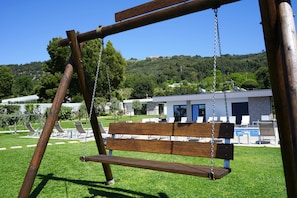 A wooden swing set in a grassy area with a modern building in the background. - Tiny Home 'Casa Azul' with Sea View, Shared Pool and Wi-Fi (Cristelo)
