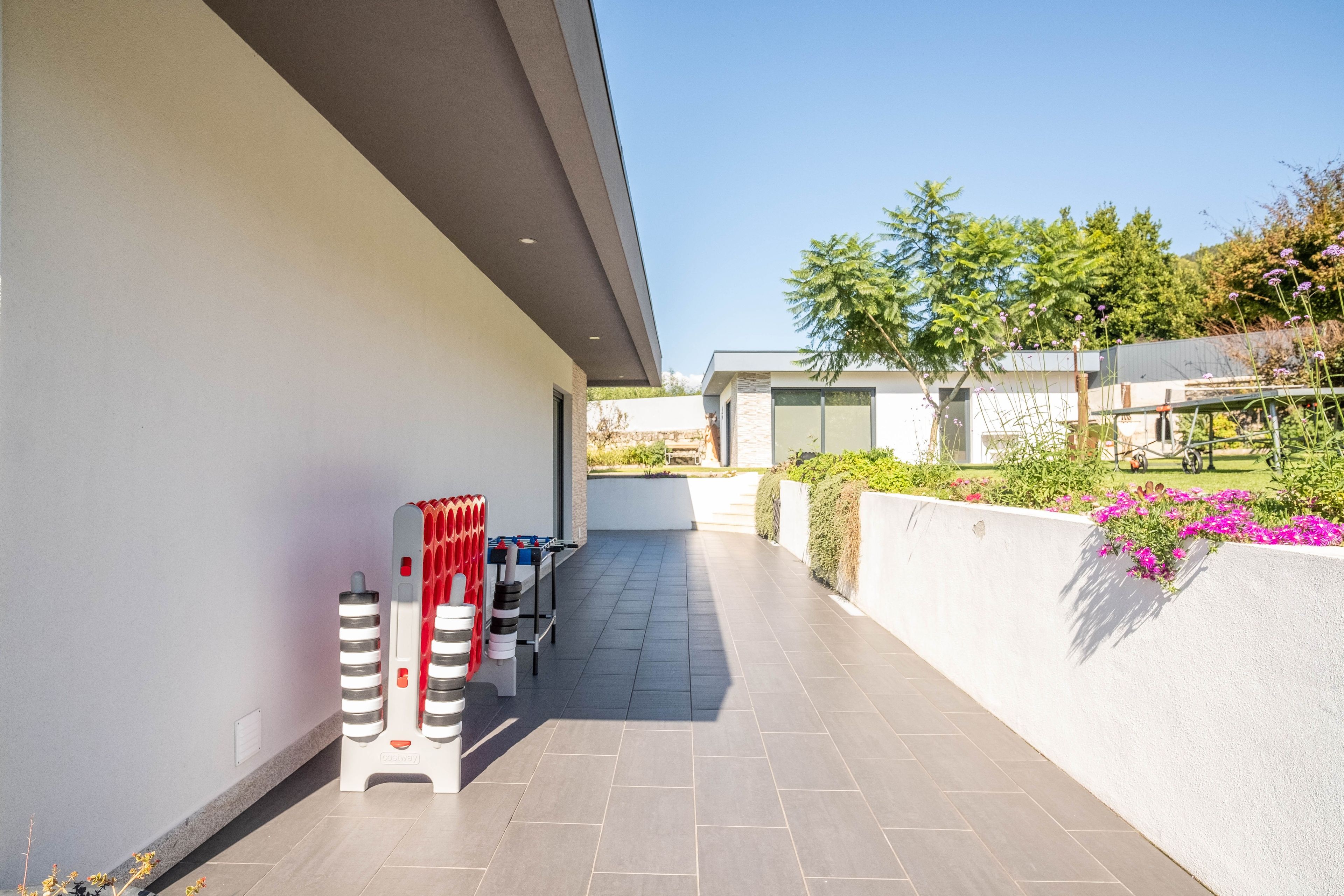 A modern outdoor area with a foosball table, a wall-mounted air conditioning unit, and a garden with flowers and shrubs.