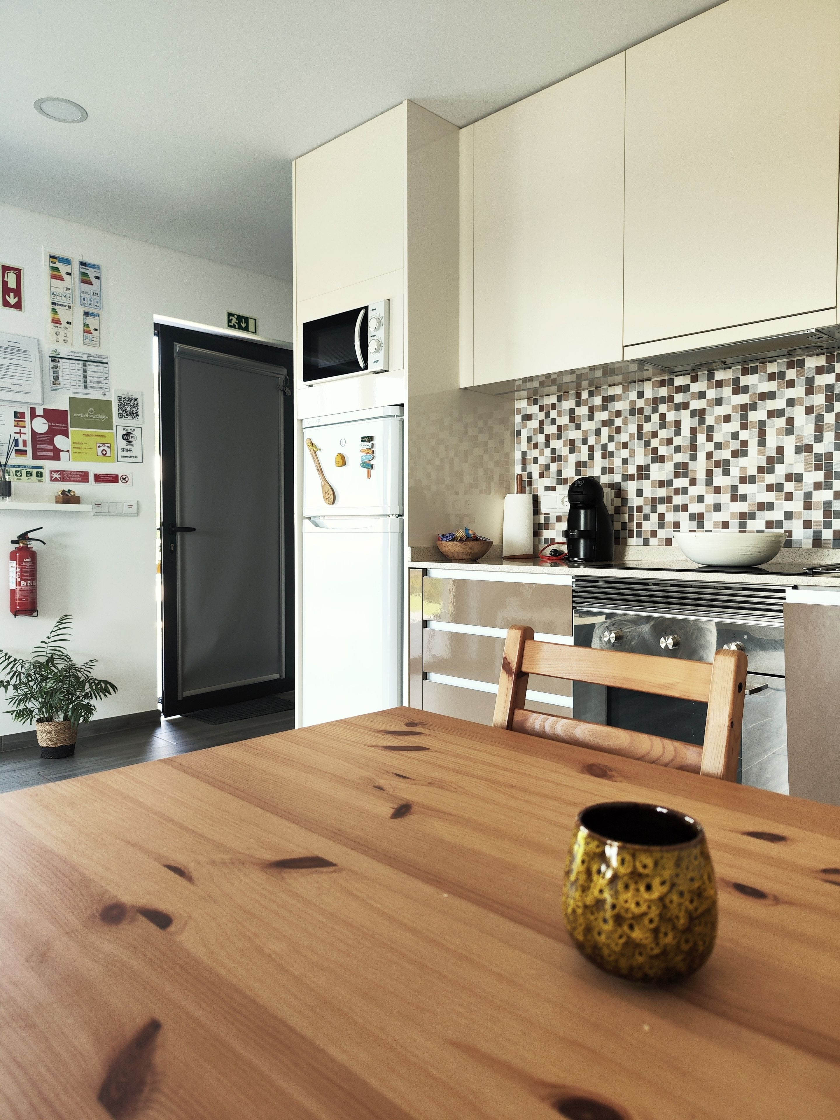 A modern kitchen with white cabinets, a microwave, and a refrigerator. There is a wooden dining table with a bowl and a mug.