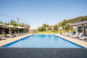 Una piscina con sillas de descanso y sombrillas, rodeada de vegetación y colinas.