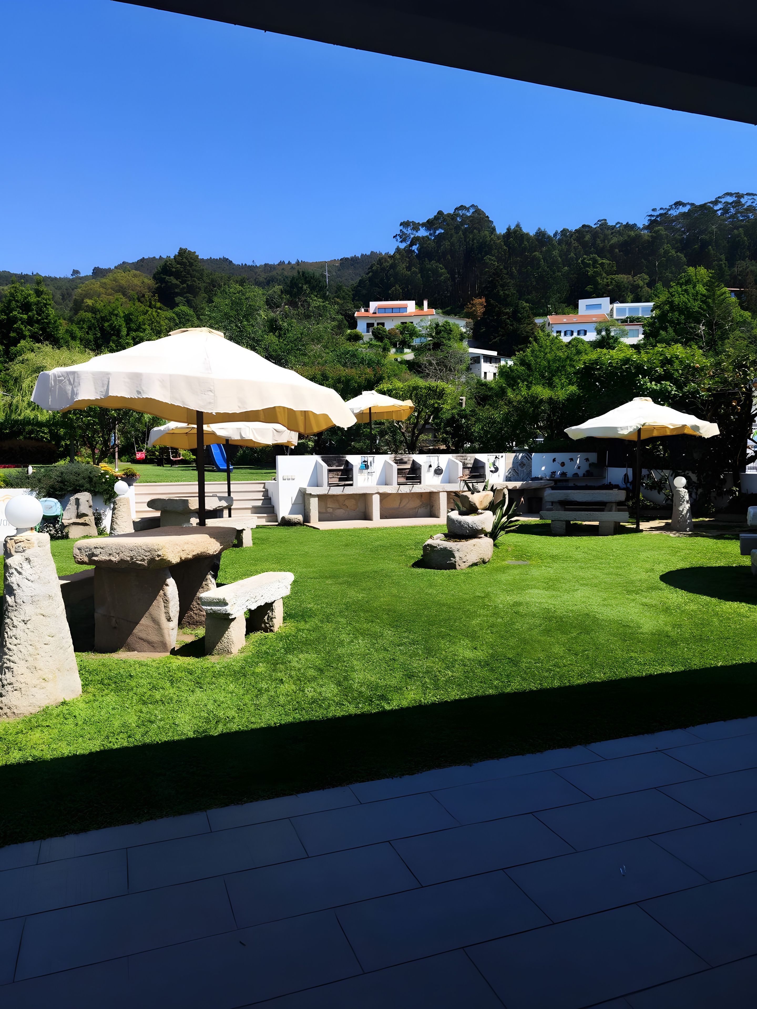 Ein Terrassenbereich im Freien mit Steintischen und -bänken, Sonnenschirmen und einem Grill.
