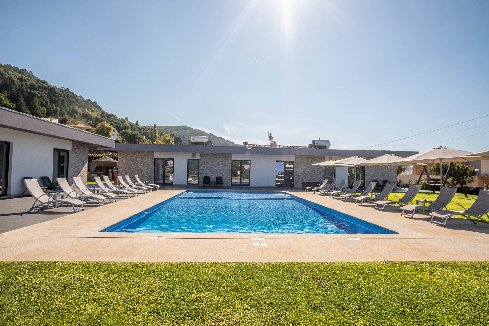 A swimming pool with lounge chairs and umbrellas, surrounded by a building with large windows and a view of a hilly landscape.