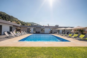 A swimming pool with lounge chairs and umbrellas, surrounded by a building with large windows and a view of a hilly landscape. - Tiny Home 'Casa Amarela' with Sea View, Shared Pool and Wi-Fi (Cristelo)