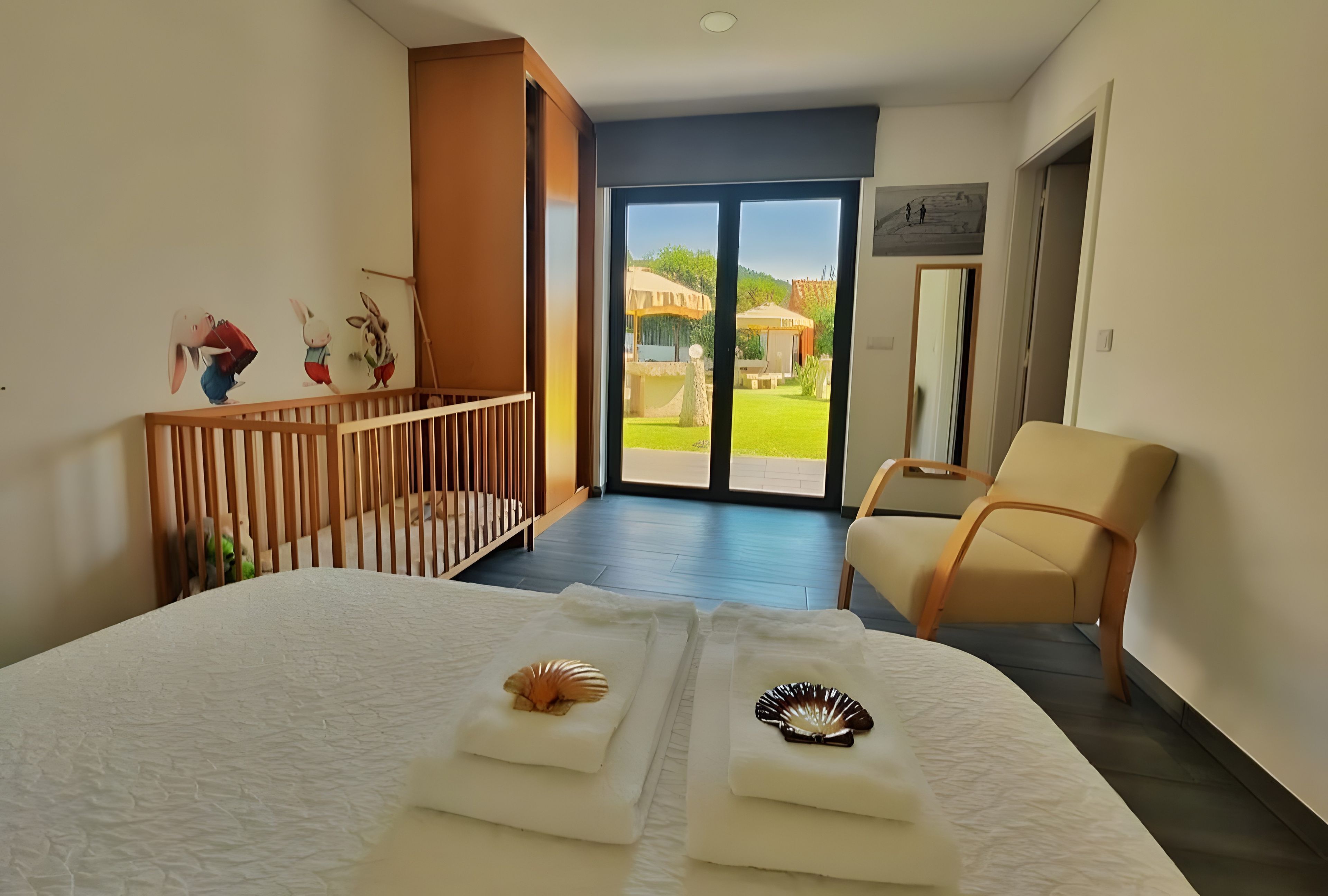 A hotel room with a bed, a chair, a crib, and a view of the outdoors.