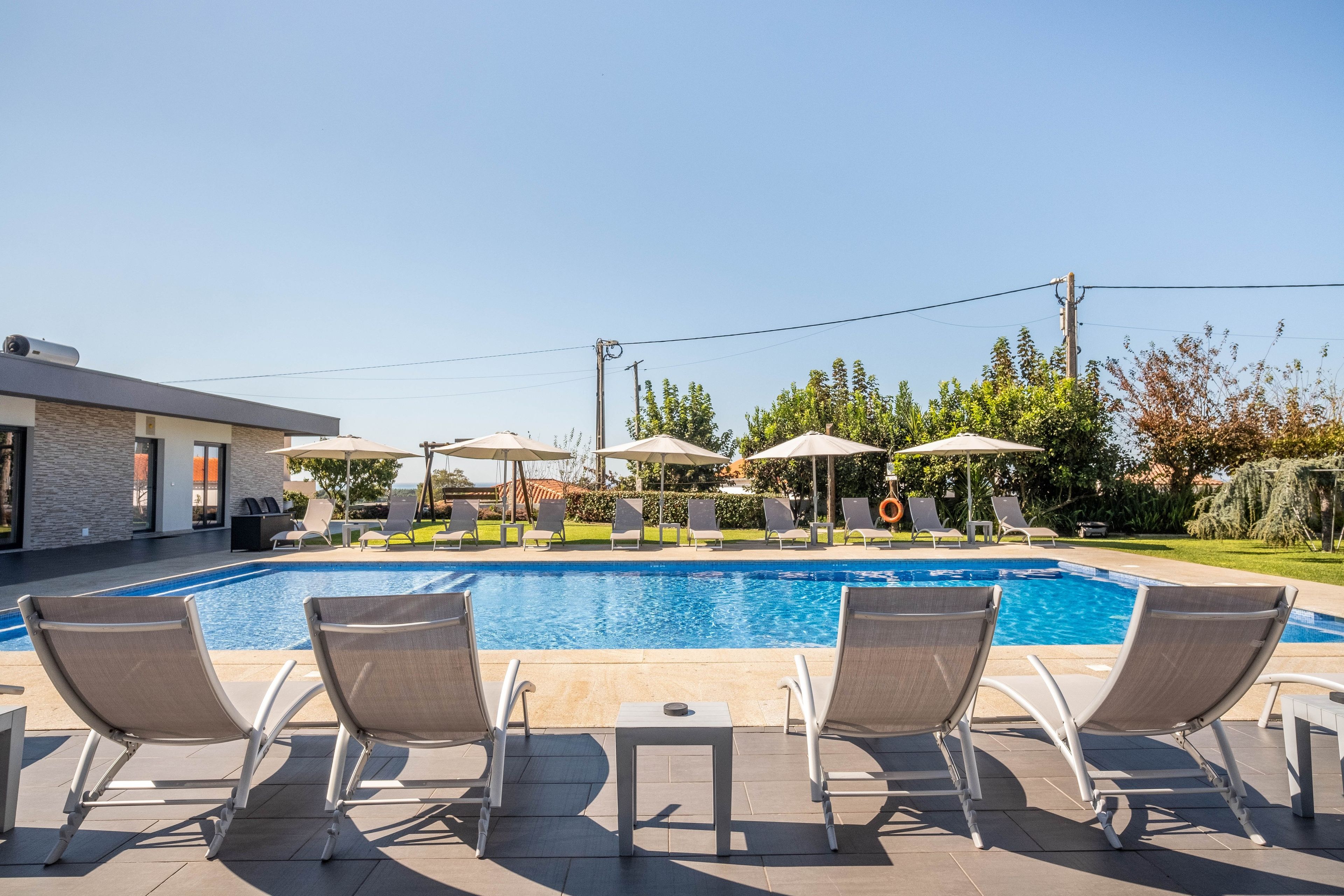 A swimming pool with lounge chairs and umbrellas, surrounded by greenery and a building.