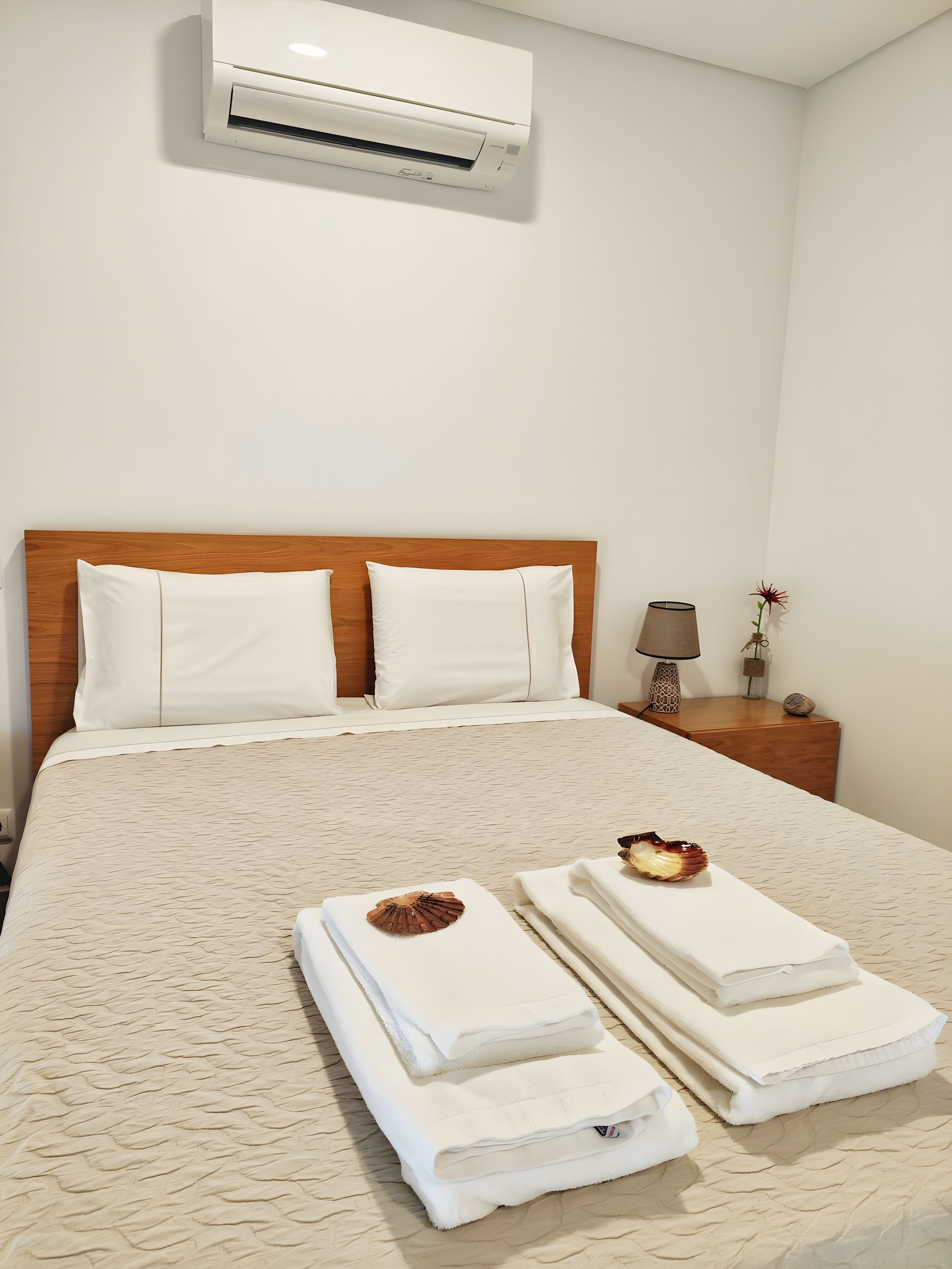 A neatly made bed with white linens and a textured beige bedspread, a wooden headboard, a bedside table with a lamp, and an air conditioning unit mounted on the wall.