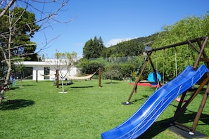 A playground with a blue slide and swings, surrounded by a grassy area and trees. - Tiny Home 'Casa Amarela' with Sea View, Shared Pool and Wi-Fi (Cristelo)