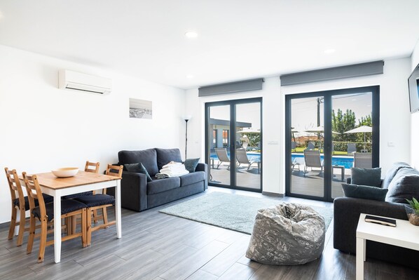 A modern living room with a dining area, a grey sofa, a bean bag, and a view of an outdoor pool area. - Tiny Home 'Casa Amarela' with Sea View, Shared Pool and Wi-Fi (Cristelo)