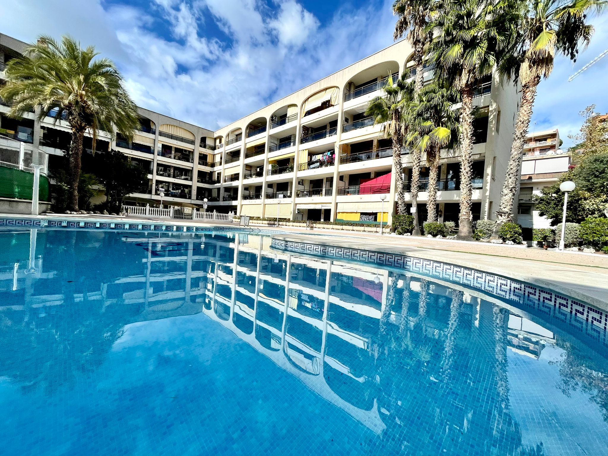 A swimming pool with a clear blue water surface, surrounded by a multi-story building with balconies, palm trees, and a clear sky.