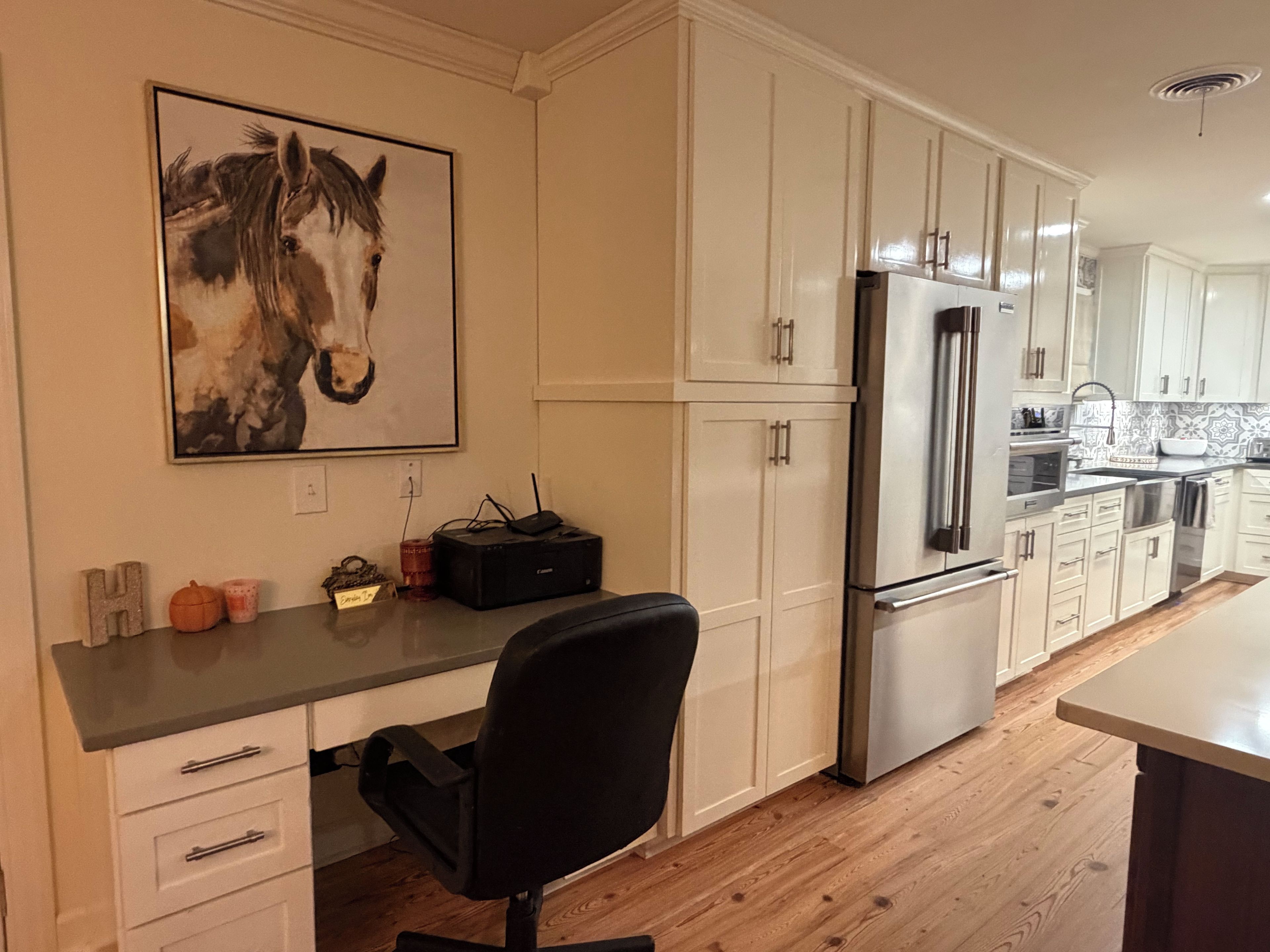 A kitchen with white cabinets, a stainless steel refrigerator, a black office chair, and a horse painting on the wall.