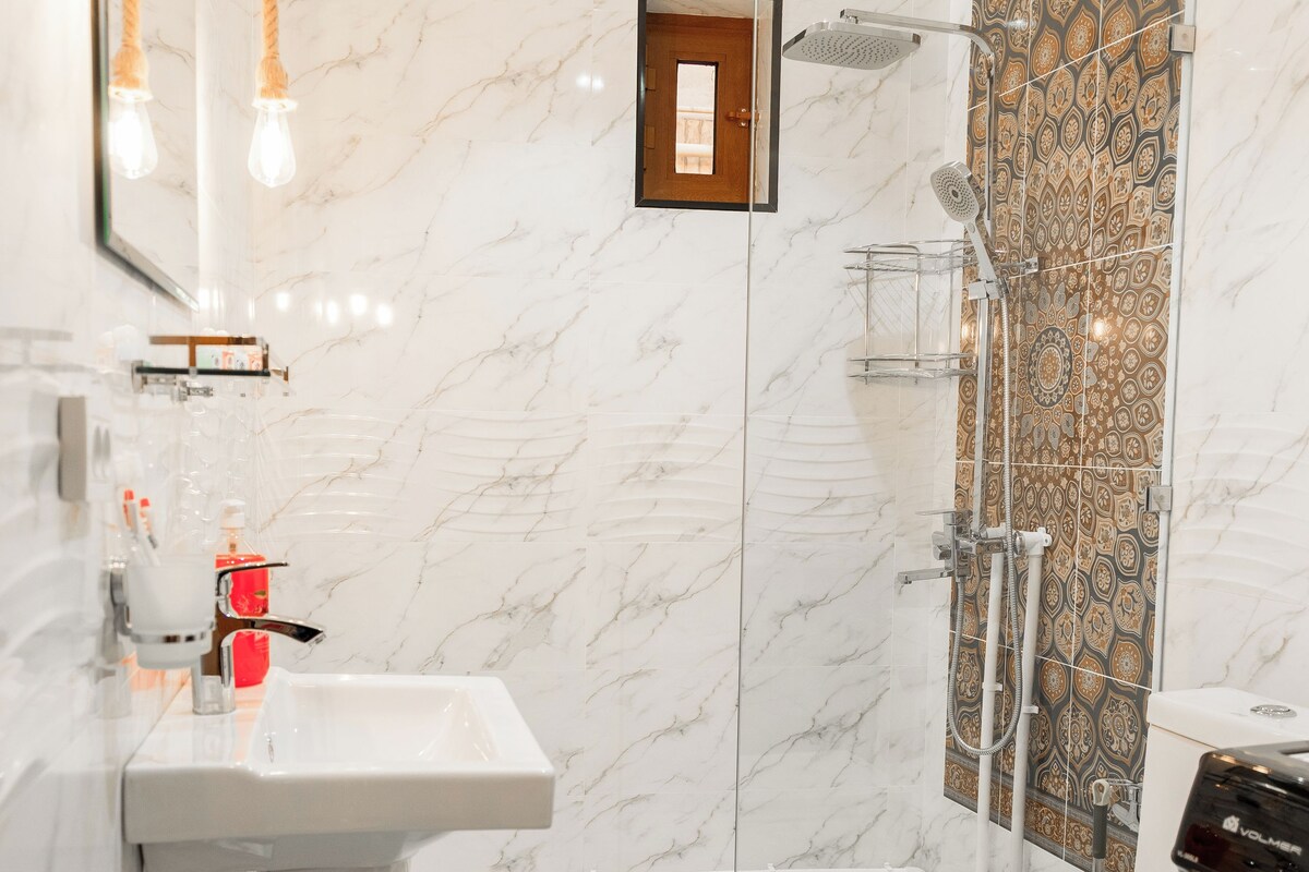 Et moderne badeværelse med klinkegulv af marmor, en hvid vask, en glasbrusekabine og et toilet.
