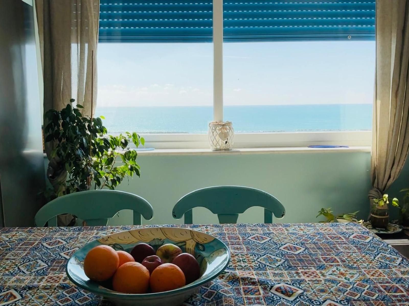Une table à manger avec un saladier de fruits, deux chaises, une plante en pot et une vue sur la mer par une fenêtre aux volets bleus.