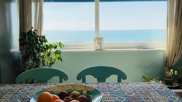 A dining table with a bowl of fruit, two chairs, a potted plant, and a view of the sea through a window with blue shutters.