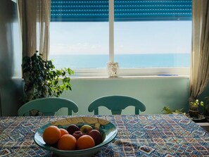 Une table à manger avec un saladier de fruits, deux chaises, une plante en pot et une vue sur la mer par une fenêtre aux volets bleus.