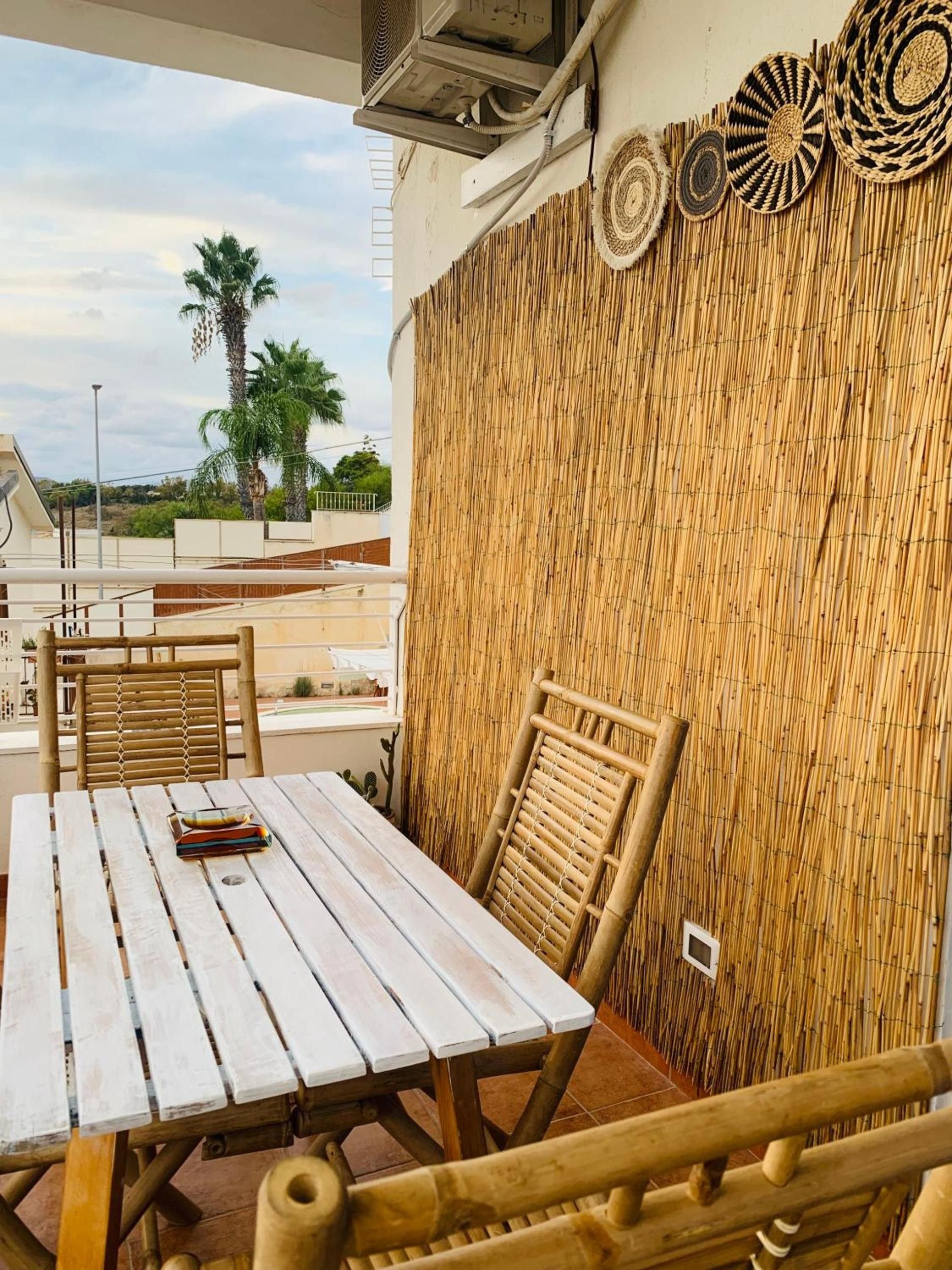 Une table et des chaises en bois, disposées dans un espace extérieur abrité, avec vue sur des palmiers et un bâtiment en arrière-plan.