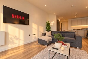 Een moderne woonkamer met een grijze bank, een salontafel met een afstandsbediening, een brandende kaars en een potplant.