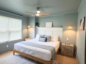 Une chambre à coucher avec un lit, des tables de chevet, un ventilateur de plafond et une fenêtre avec des stores.