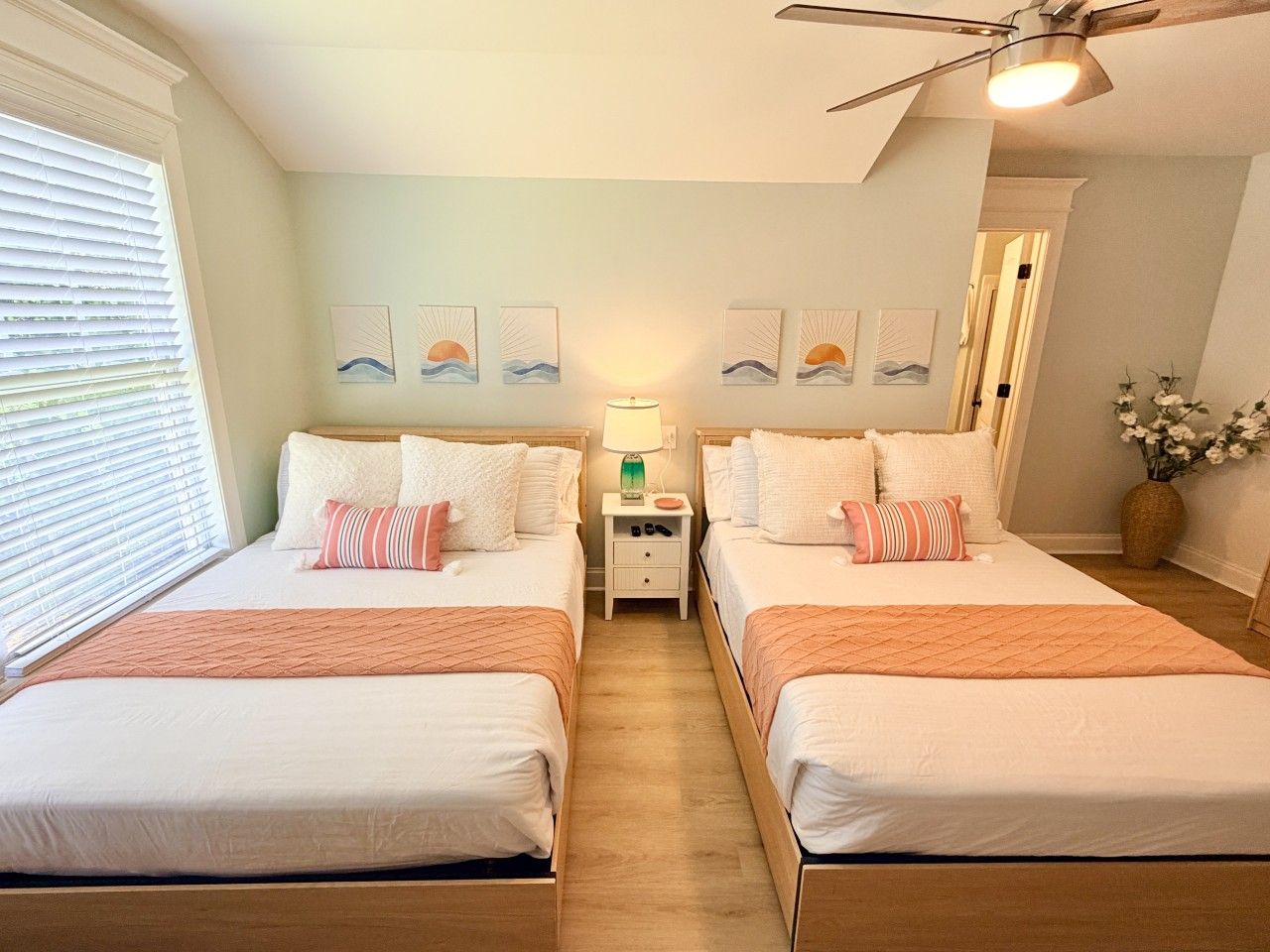 A hotel room with two beds, a nightstand, a ceiling fan, and artwork on the walls.