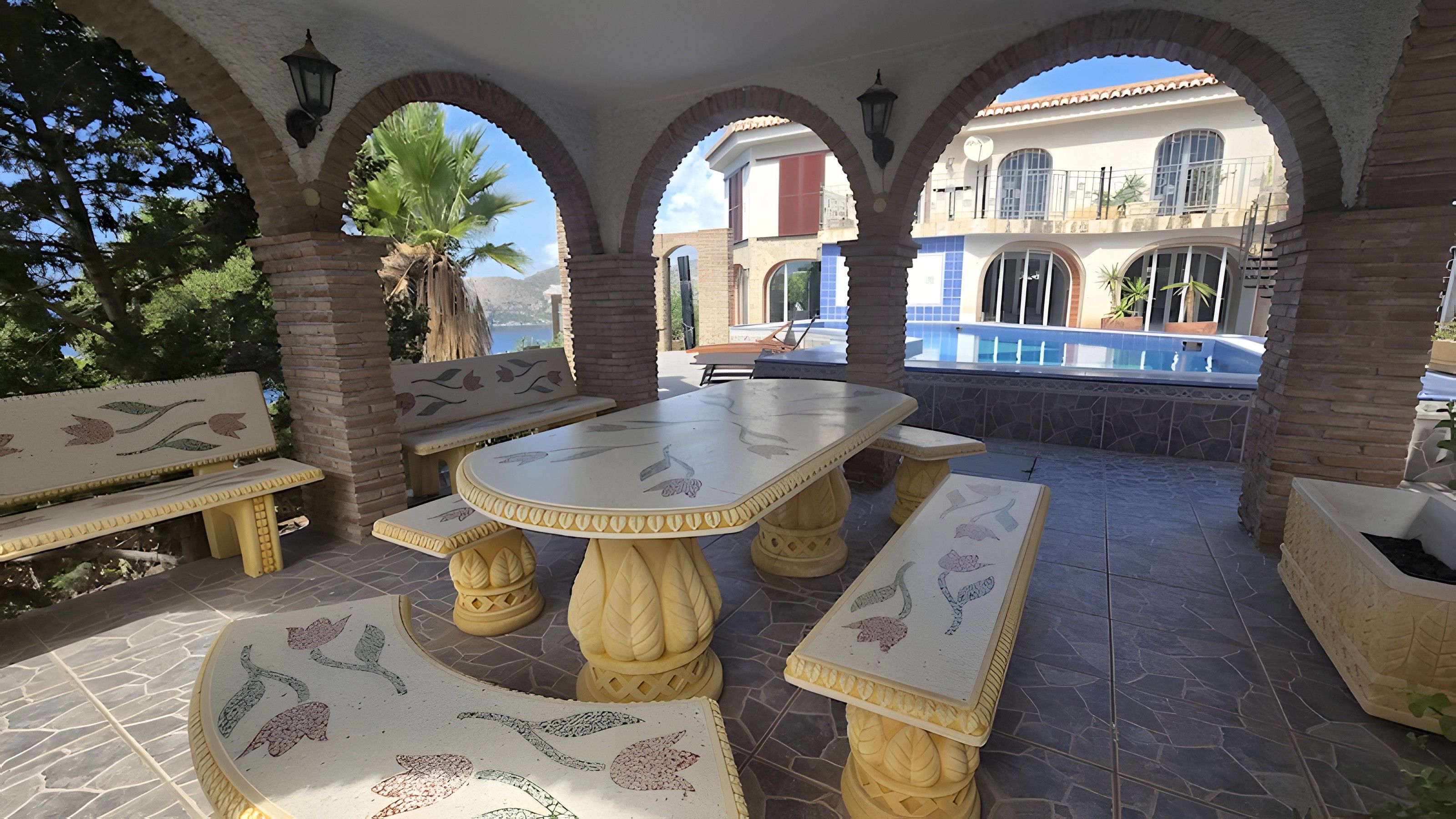 Une terrasse extérieure avec une table et des bancs, entourée d’arcades et avec une piscine en arrière-plan.