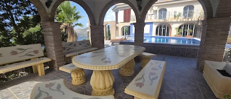 Un patio exterior con una mesa y bancos, rodeado de arcos y una piscina al fondo.