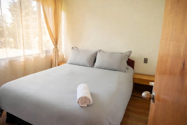 A bed with a rolled white towel, a wooden nightstand, and a wall-mounted light switch.