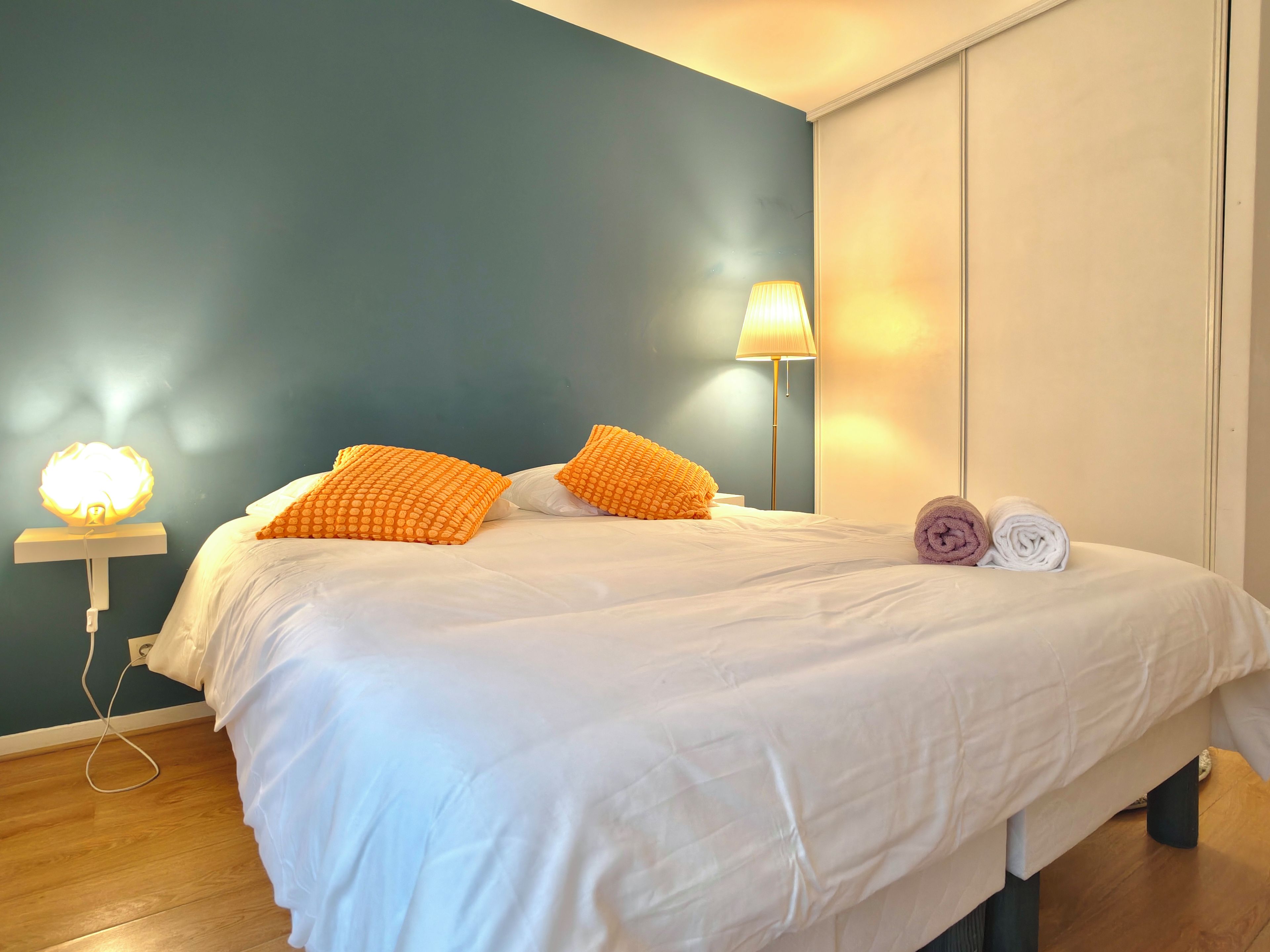A neatly made bed with white bedding, two orange pillows, and a rolled towel on the side.