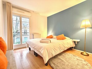 Quarto com cama, mesa de cabeceira, abajur e vista do exterior pela janela.