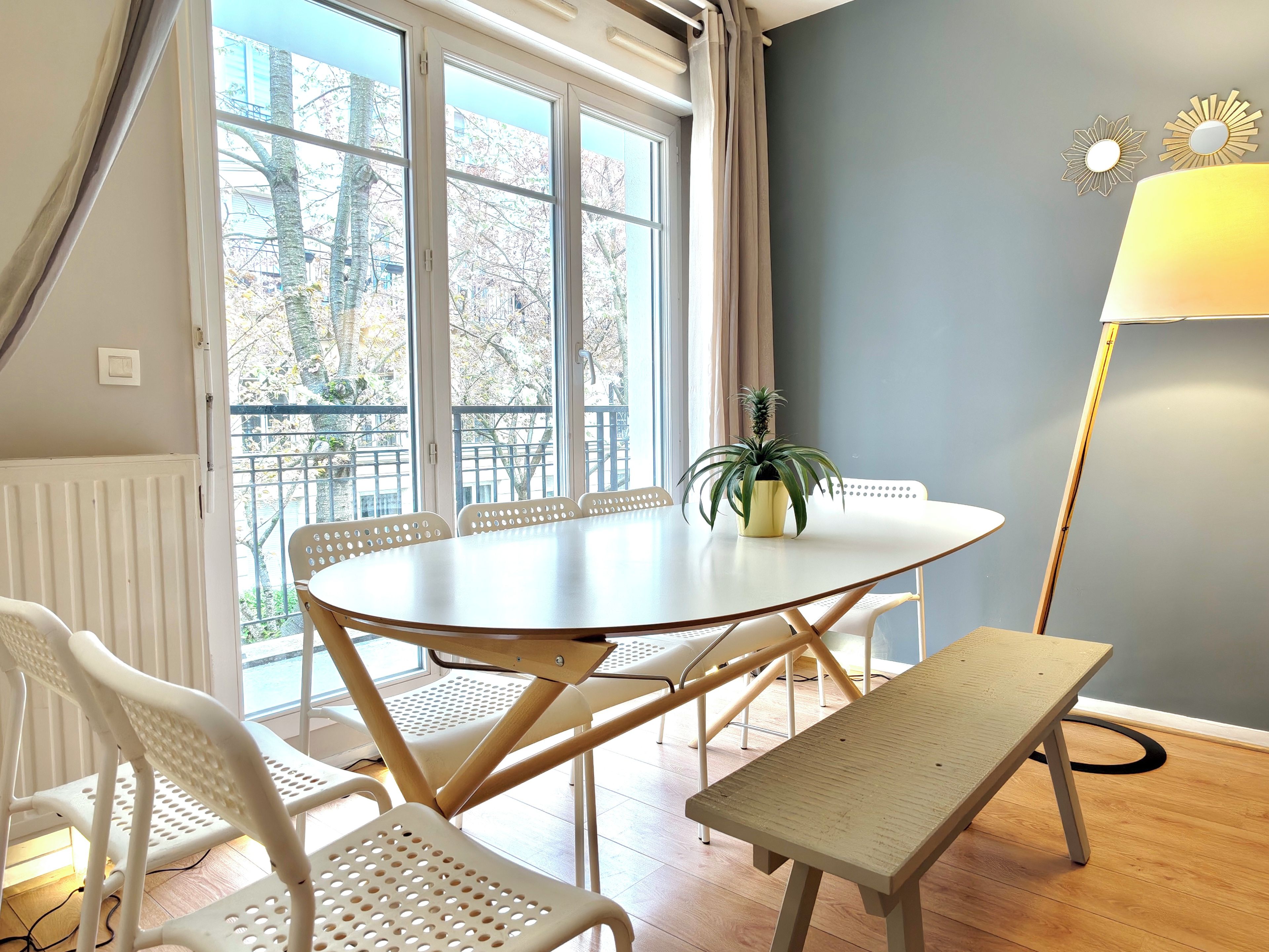 A room with a round table, white chairs, a bench, a potted plant, a lamp, and a view through large windows.