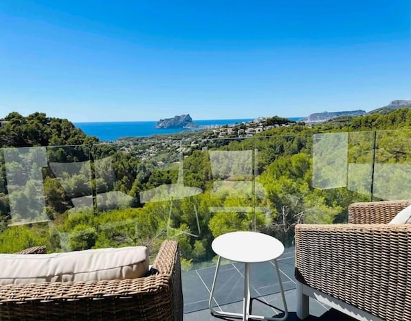 A balcony with wicker furniture and a view of the sea and hills.