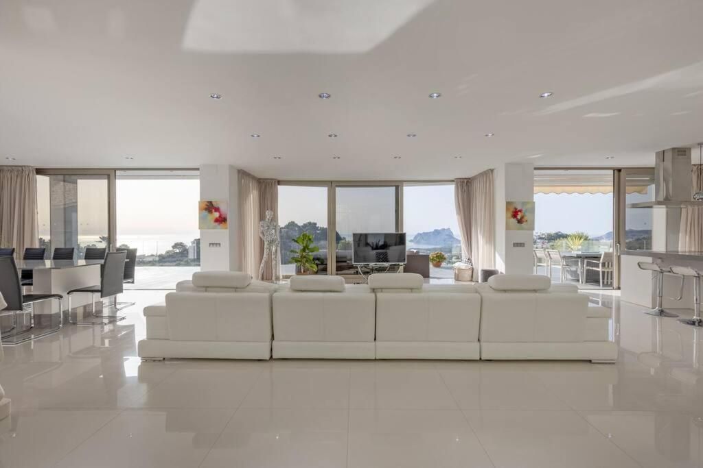 A modern living room with a white sofa set, a dining area with a glass table, and a large window offering a view of the sea.