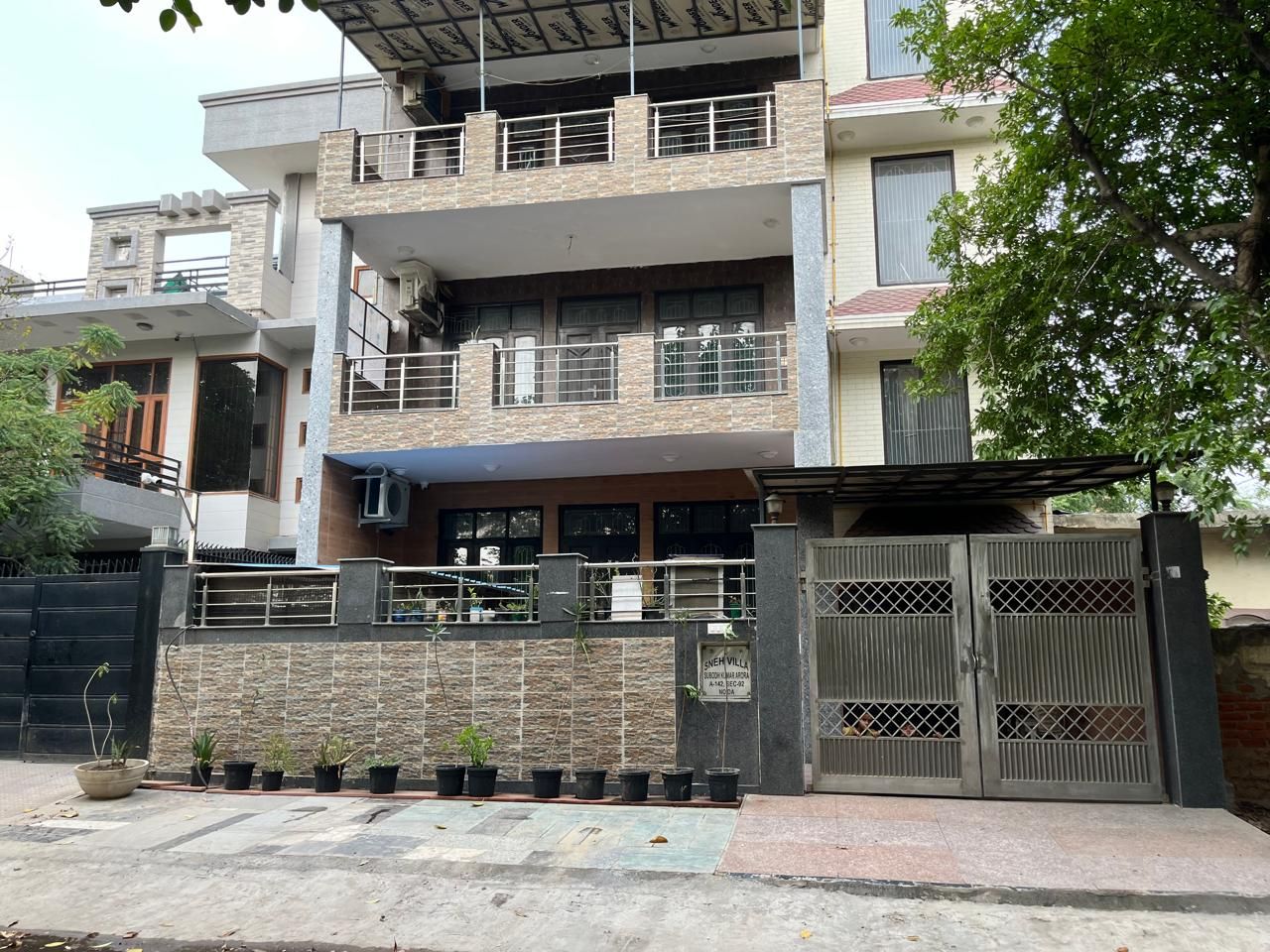 Un edificio residencial de varios pisos con entrada protegida por un portón, un balcón en el nivel superior y un muro de piedra con plantas en macetas.
