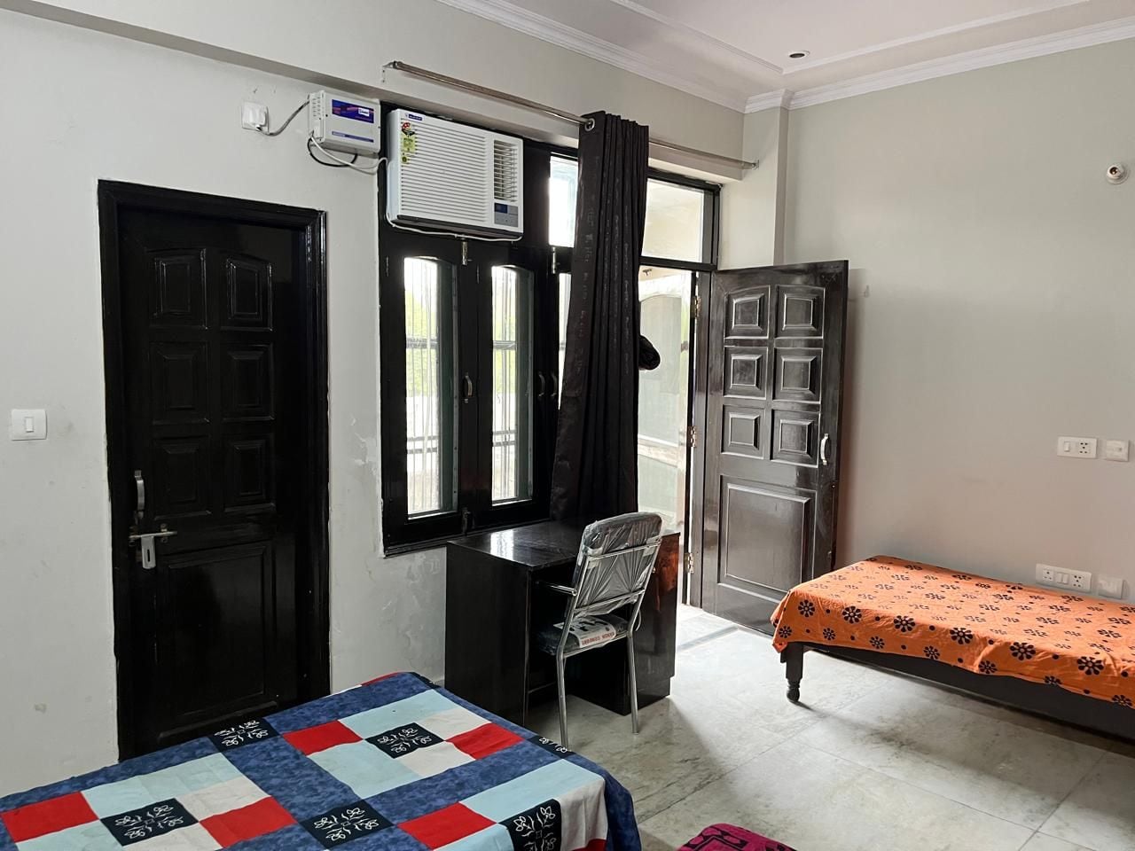 Una habitación con cama, escritorio y silla. Cuenta con una ventana con cortinas, una puerta y un equipo de aire acondicionado.