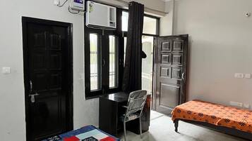 Una habitación con cama, escritorio y silla. Cuenta con una ventana con cortinas, una puerta y un equipo de aire acondicionado.