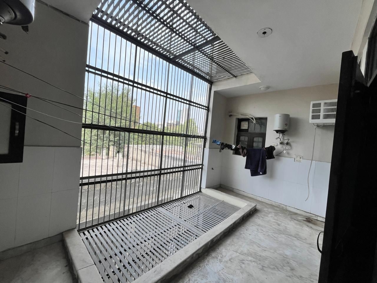 Una habitación con una ventana con barrotes, un banco pequeño y un equipo de aire acondicionado fijado a la pared.