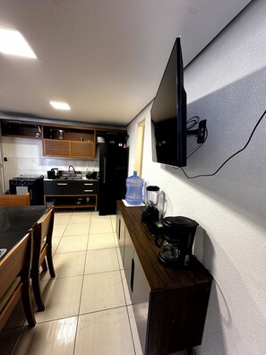 Una cocina compacta con una mesa de comedor, sillas, un refrigerador, dispensador de agua, cafetera y un televisor de pantalla plana montado en la pared.